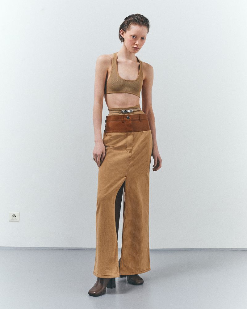 Woman in a tan halter top and brown skirt stands in a studio with a neutral expression, showcasing a modern fashion look.