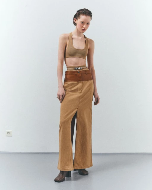 Woman in a tan halter top and brown skirt stands in a studio with a neutral expression, showcasing a modern fashion look.