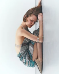 Model wearing a textured knit dress and silver chain jewelry seated against a pale wall for DUEL magazine.