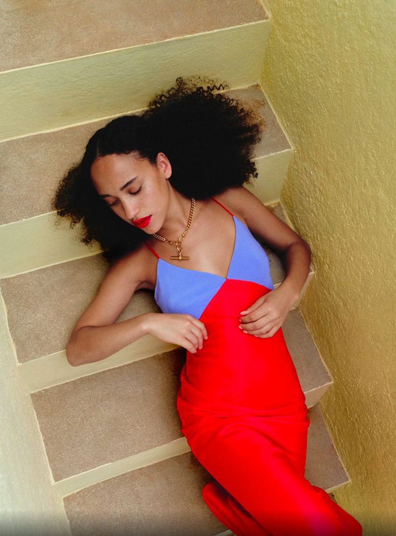 Woman in red and blue dress lies on yellow stairs, wearing a gold necklace with red lipstick and dark curly hair.