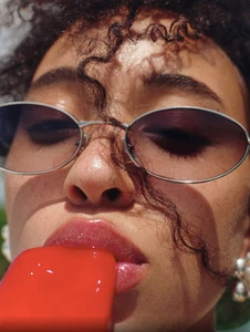 Close-up portrait of a woman wearing thin metal sunglasses and eating a bright red popsicle for Cosmopolitan UK beauty editorial.