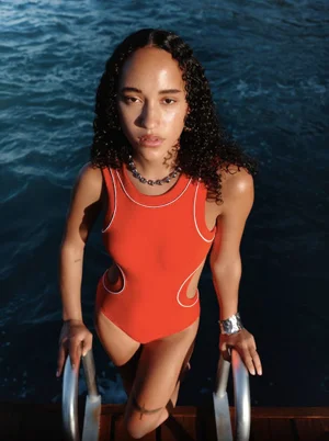 Woman in a red swimsuit standing on a wooden dock with dark water in the background by Cosmopolitan UK