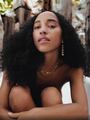 Close-up portrait of a woman with long dark curly hair wearing gold necklaces and earrings against a textured background.