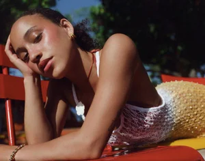 Close-up portrait of a woman resting her head on her hand outdoors wearing a white beaded top.