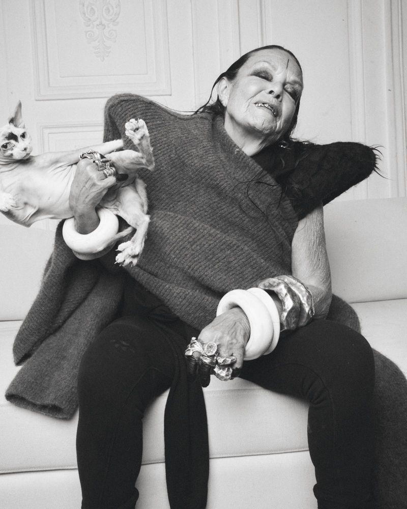 Monochrome portrait of an elderly woman holding a hairless cat, seated indoors, wearing jewelry and a dark shawl, with a decorative background.