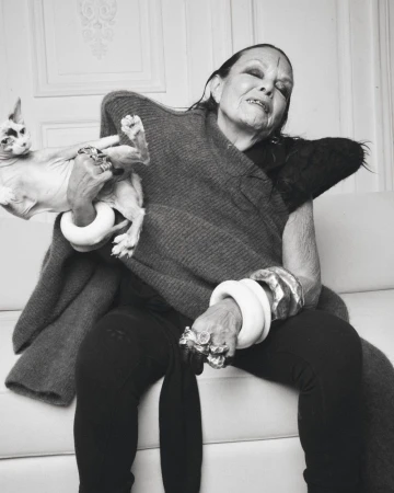 Monochrome portrait of an elderly woman holding a hairless cat, seated indoors, wearing jewelry and a dark shawl, with a decorative background.