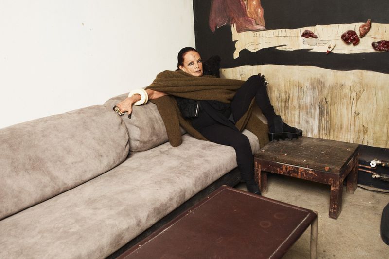 Mature woman lounges on a grey couch with an olive shawl and black clothes in a casual, artistic interior setting with expressive art.