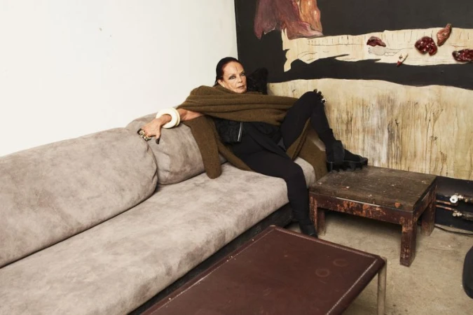 Mature woman lounges on a grey couch with an olive shawl and black clothes in a casual, artistic interior setting with expressive art.