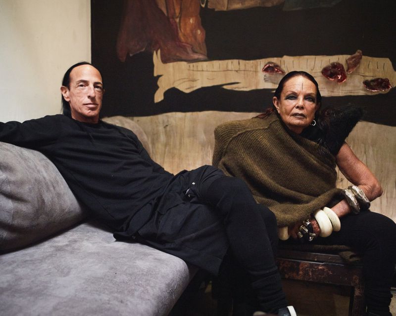 Candid shot of Rick Owens and Michele Lamy sitting indoors on a couch, posed in front of a piece of art. Neutral tones.