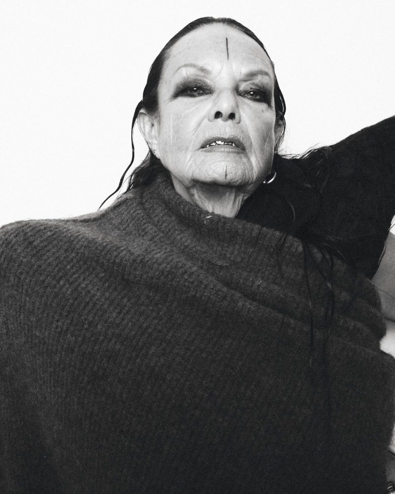 Monochrome portrait of an older woman with dark eye makeup and face markings, wrapped in a dark shawl against a white backdrop.