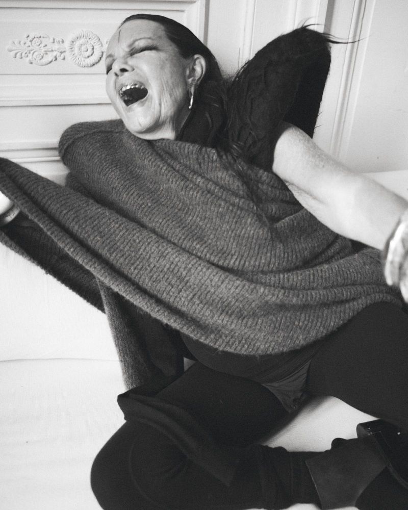 Black and white portrait of a mature woman with her mouth open wearing a sweater and boots in an indoor setting.
