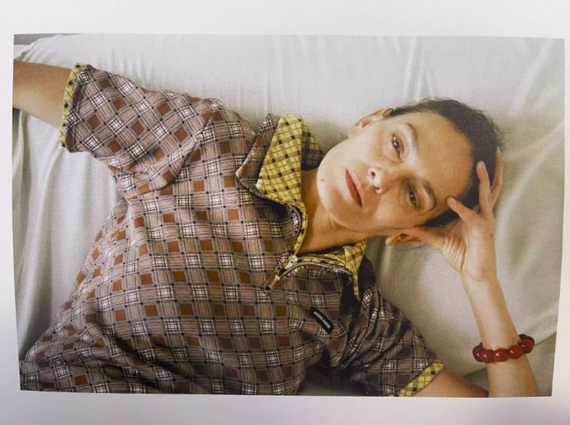 Woman in patterned pajamas reclines on white sheets, resting her head on her hand and gazing at the camera, red bracelet visible.