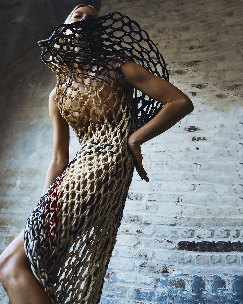 A slender adult female poses in a net-like dress against a distressed brick wall, showcasing a fashion editorial style and neutral color palette.
