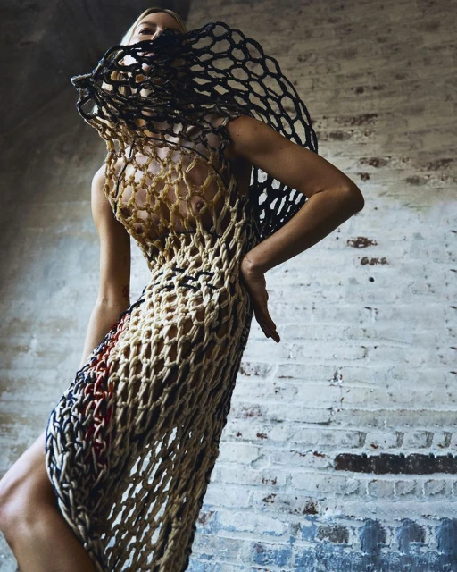 A slender adult female poses in a net-like dress against a distressed brick wall, showcasing a fashion editorial style and neutral color palette.