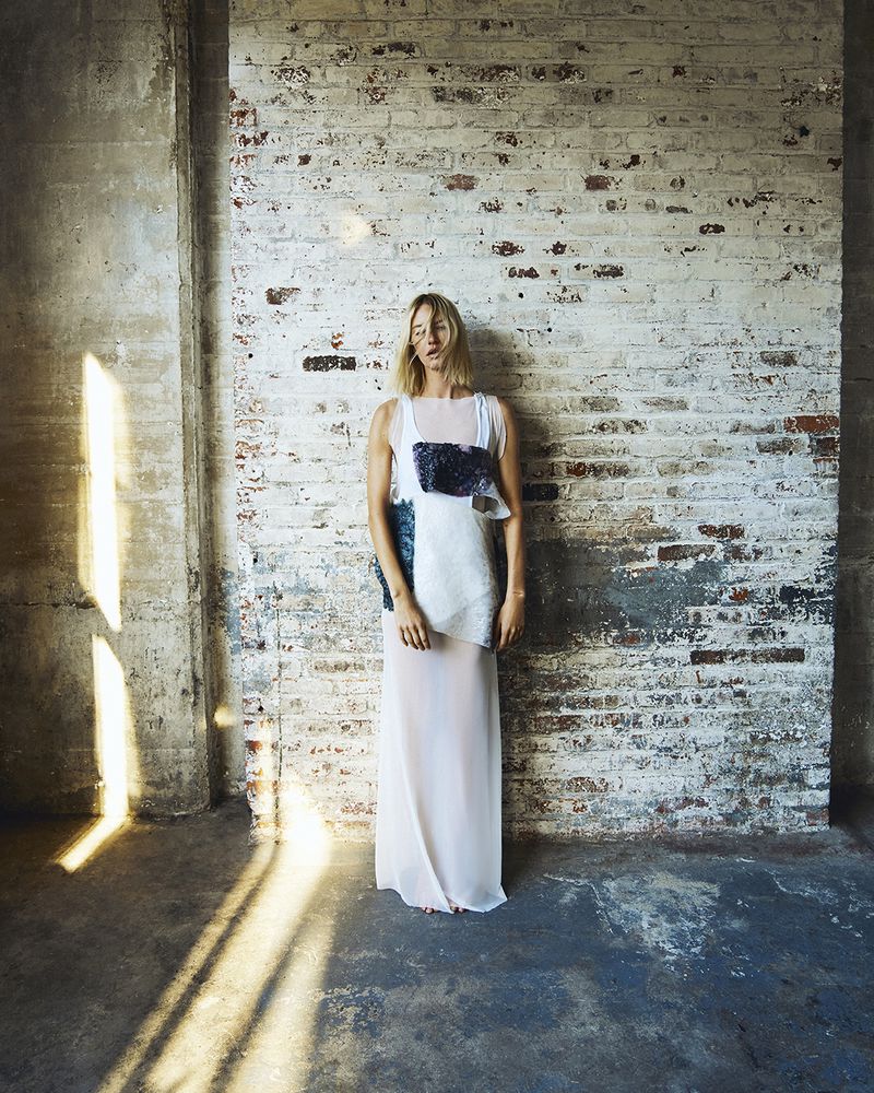 A woman stands in a sheer white dress with a blue-and-white top against a distressed brick wall, with sunlight and shadows.