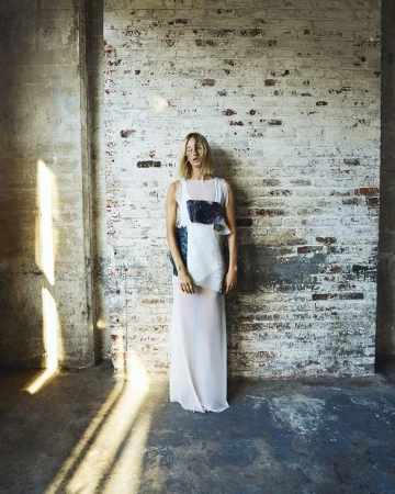 A woman stands in a sheer white dress with a blue-and-white top against a distressed brick wall, with sunlight and shadows.