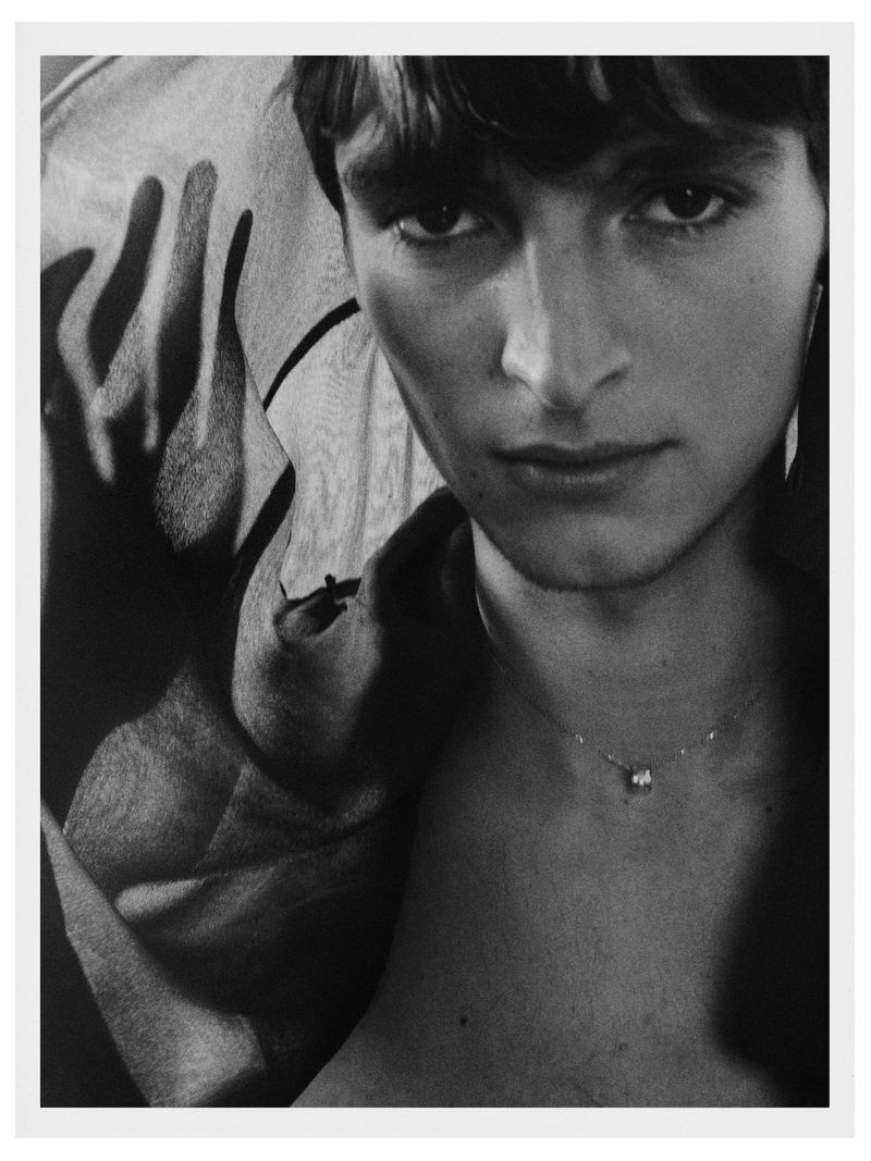 Black and white portrait of a young adult male with a bare chest and a necklace, posing in front of patterned fabric backdrop.
