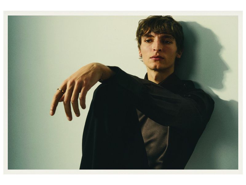 Portrait of a young adult male in a black shirt leaning against a light blue wall with his hand raised, fingers spread, and wearing a ring.
