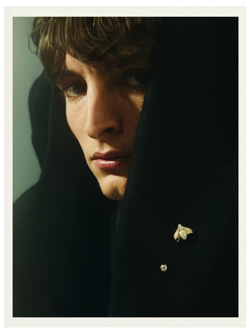 Portrait of a young adult man with dark hair partially concealed by black fabric, accessorized with a bee brooch and stud earring.