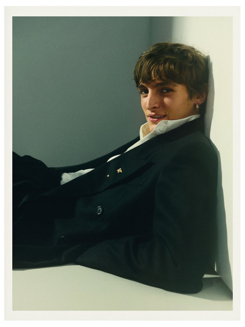 Portrait of a young adult man with dark hair, wearing a black blazer and a white shirt, leaning against a white wall in a photo studio.