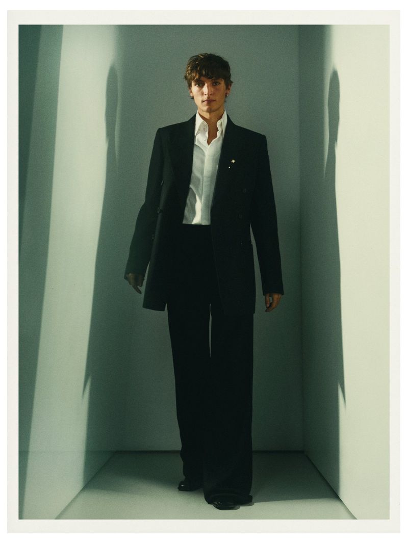 A stylish model in a black suit and white shirt walks in a photo studio with clean lines, soft lighting, and a neutral color palette.