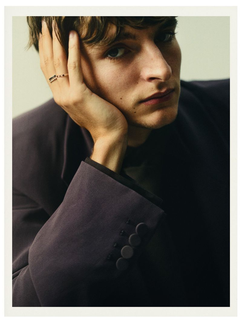 Portrait of a young adult male with dark hair, wearing a dark purple blazer, his hand is near his face, posing indoors.