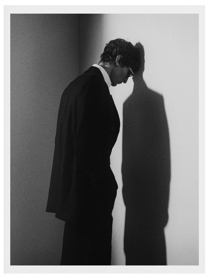 Monochrome portrait of a man in a suit standing next to a wall with his shadow projected on it in a photo studio.