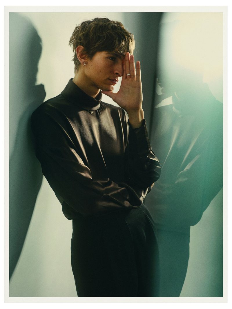Portrait of a young adult man with hand up near his face in an indoor setting, wearing a black turtleneck and pants. 