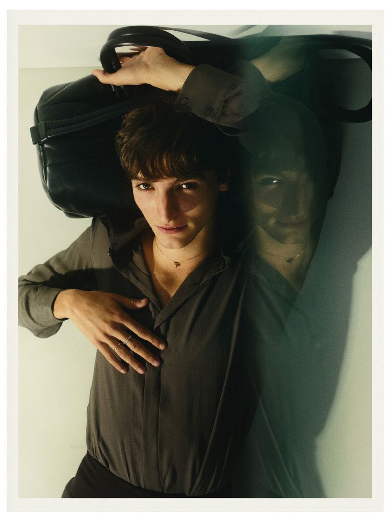 Adult laying indoors wearing buttoned shirt, pants, and necklace holding a large black leather bag with a blurry face reflection.