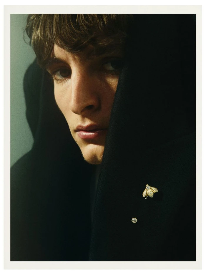 Close-up portrait of male model Vassili Schneider wearing a dark hooded garment adorned with a bee brooch for Silhouette Magazine.