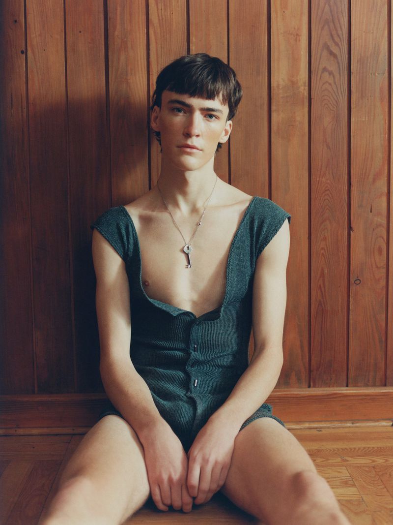 Portrait of a young adult man sitting on a wood floor in front of a wood paneled wall, wearing a one-piece and a necklace with a key pendant.