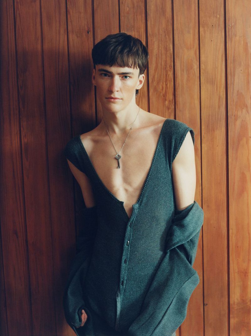 Young adult posing in a gray jumpsuit against a wood background. He wears a silver necklace with a key pendant.