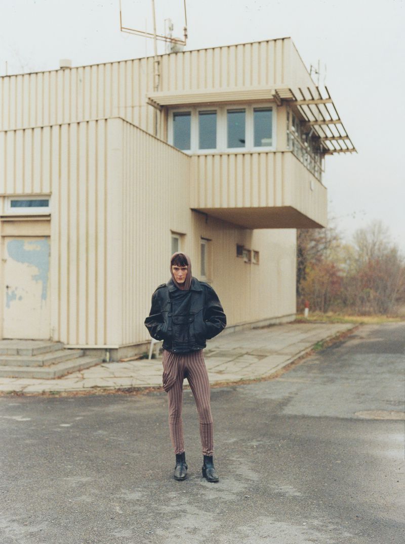 Full body shot of a young adult in a black jacket and striped pants standing in front of an industrial building on an overcast day.