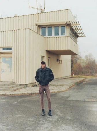 Full body shot of a young adult in a black jacket and striped pants standing in front of an industrial building on an overcast day.