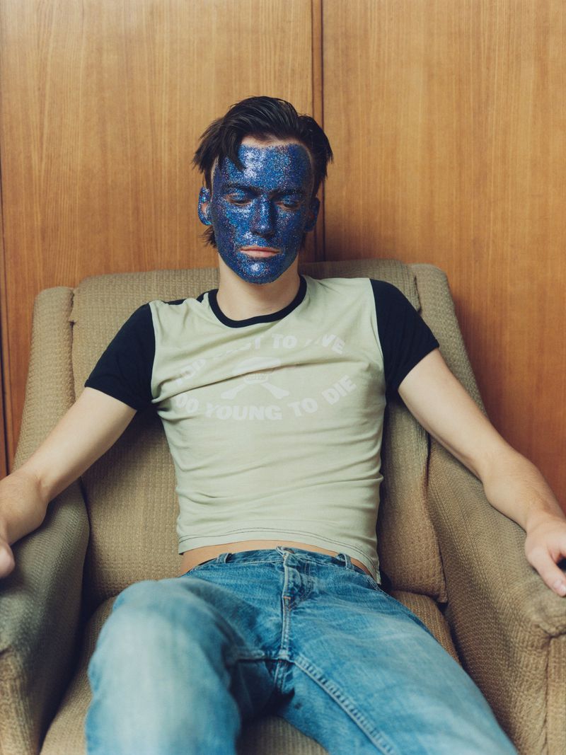 A person with blue glitter on their face sits in an armchair, wearing a graphic tee and jeans, under soft light and a simple background.