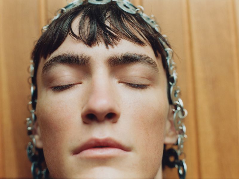 Close-up portrait of a young adult with closed eyes, adorned with a headdress made of metal rings, set against a warm wood background.