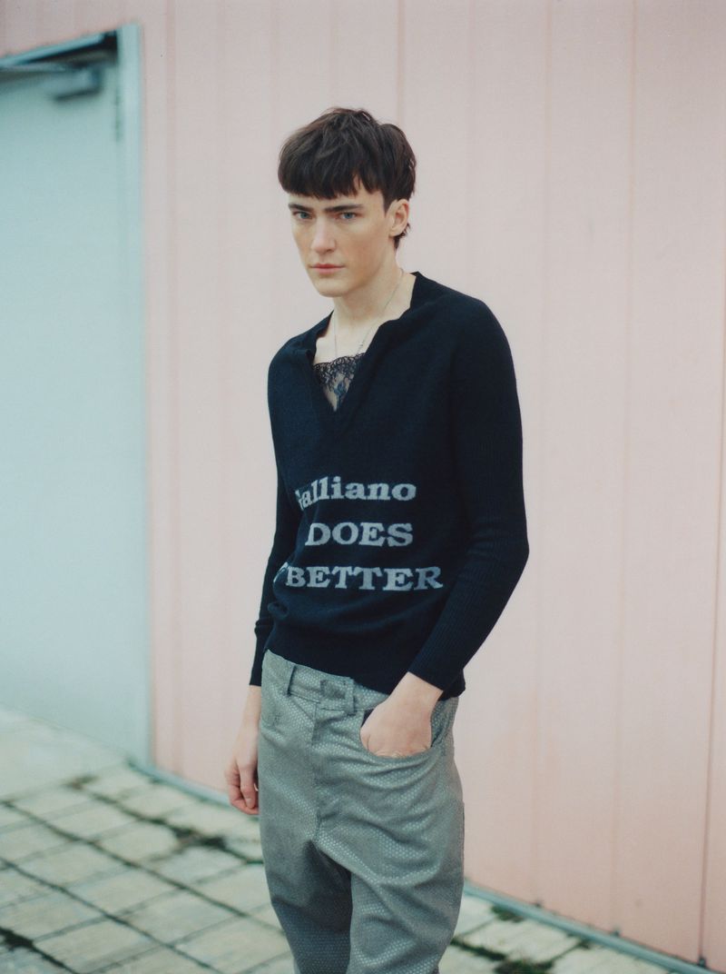Portrait of a young adult male wearing a dark sweater with text and light trousers, standing outdoors against a light pink wall.