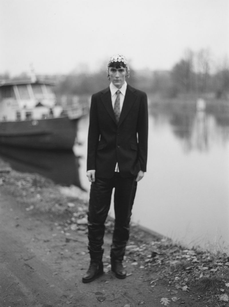 Black and white portrait of a man in a suit with floral headwear standing near a river and boat with bare trees and cloudy sky.