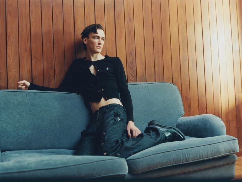 A young adult man poses on a blue couch in a black cardigan and dark denim jeans against a wood paneled wall in a fashion setting.
