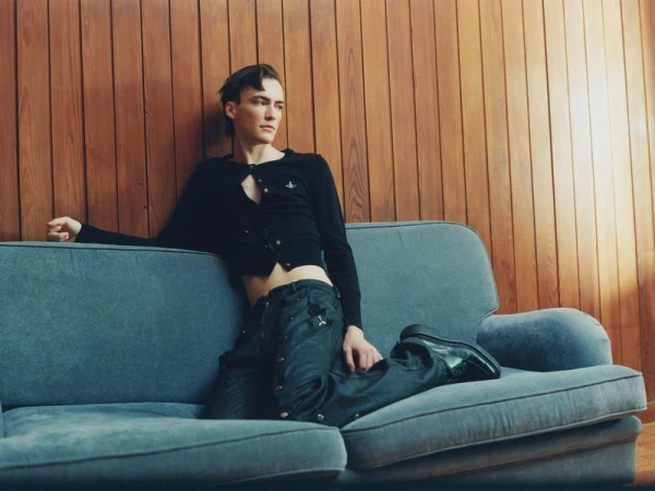 A young adult man poses on a blue couch in a black cardigan and dark denim jeans against a wood paneled wall in a fashion setting.