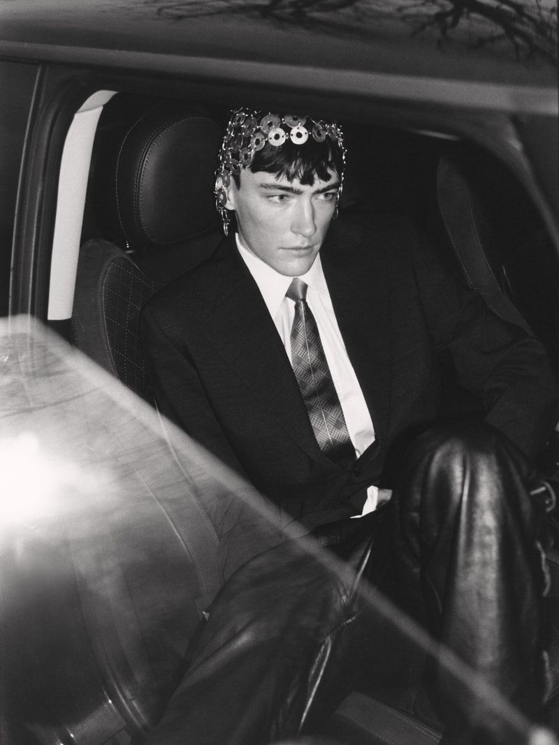 Black and white portrait of a male fashion model wearing a decorative headpiece and suit while sitting in a car interior.