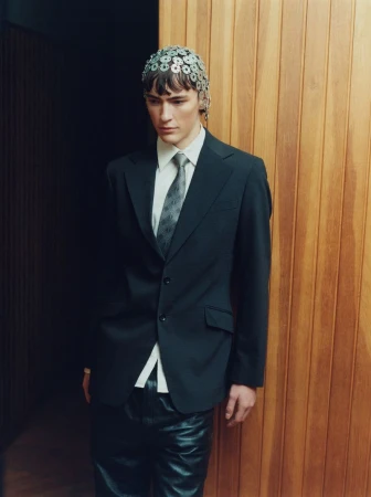 Fashion photo of a male model in a dark suit, tie, and leather pants, wearing a unique metal headpiece, posing against a wood-paneled wall.