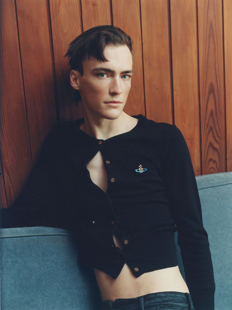 Portrait of a young adult man in a black cardigan and jeans, leaning against a blue couch and wood paneling in a studio setting.