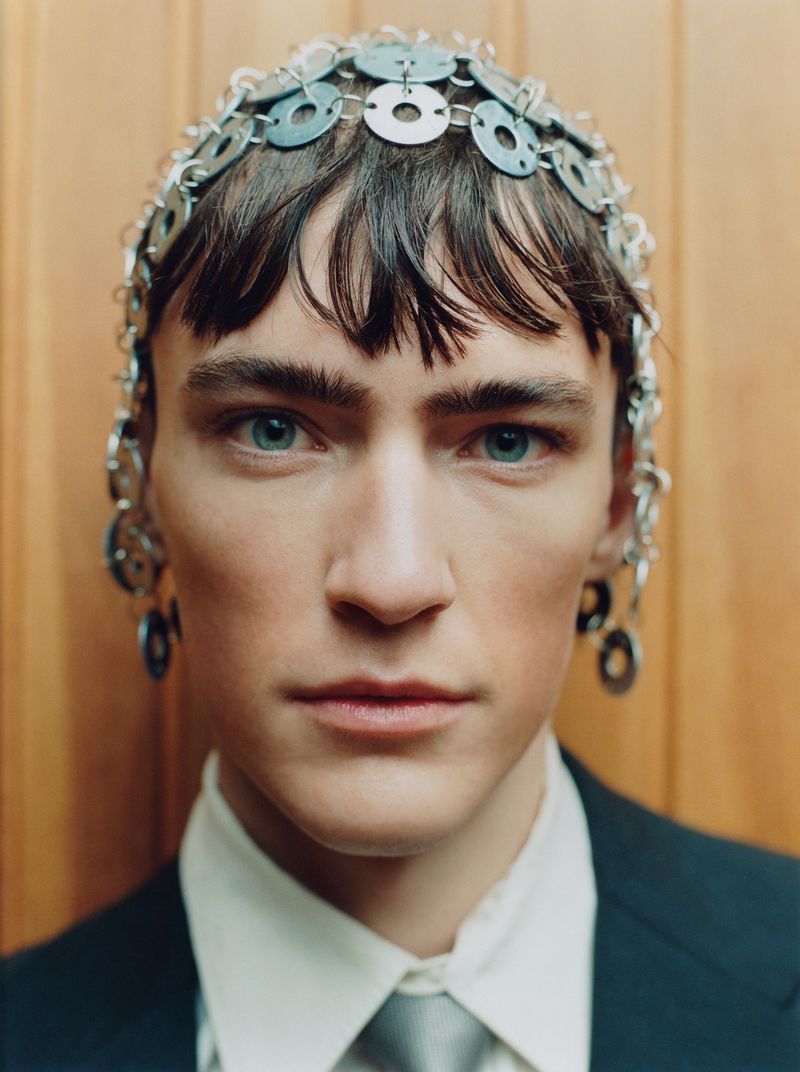 Close-up of a young adult male with a serious expression, wearing a metal headpiece and a suit, against a simple wood background.