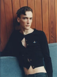 Young man wearing a black buttoned cardigan resting against a blue sofa with vertical wooden paneling behind him.