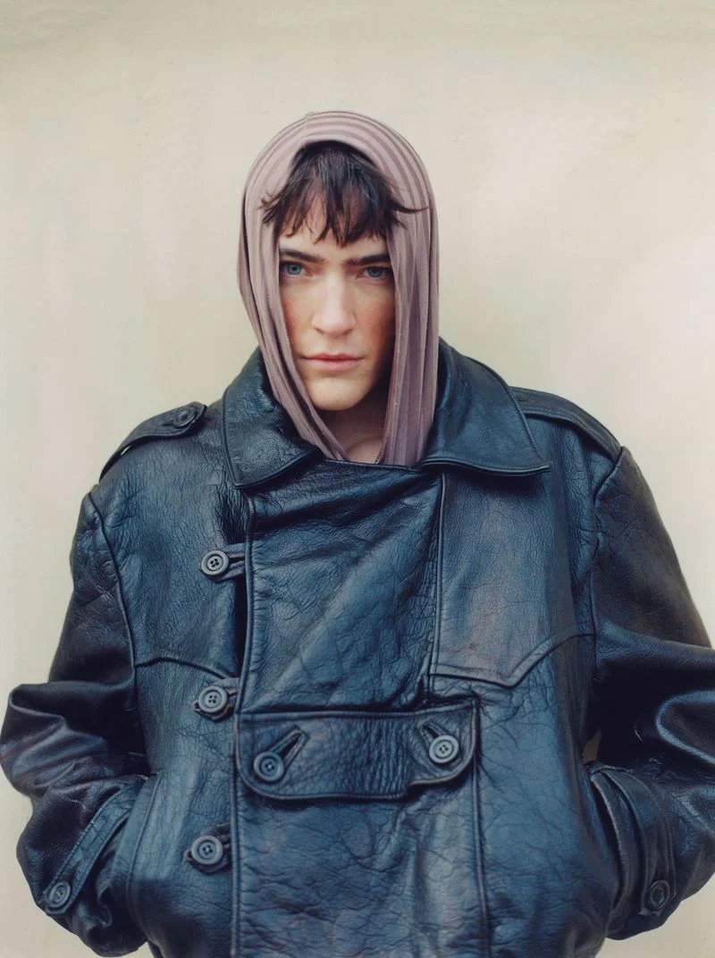 Model wearing a dark oversized leather jacket and mauve head covering against a neutral background