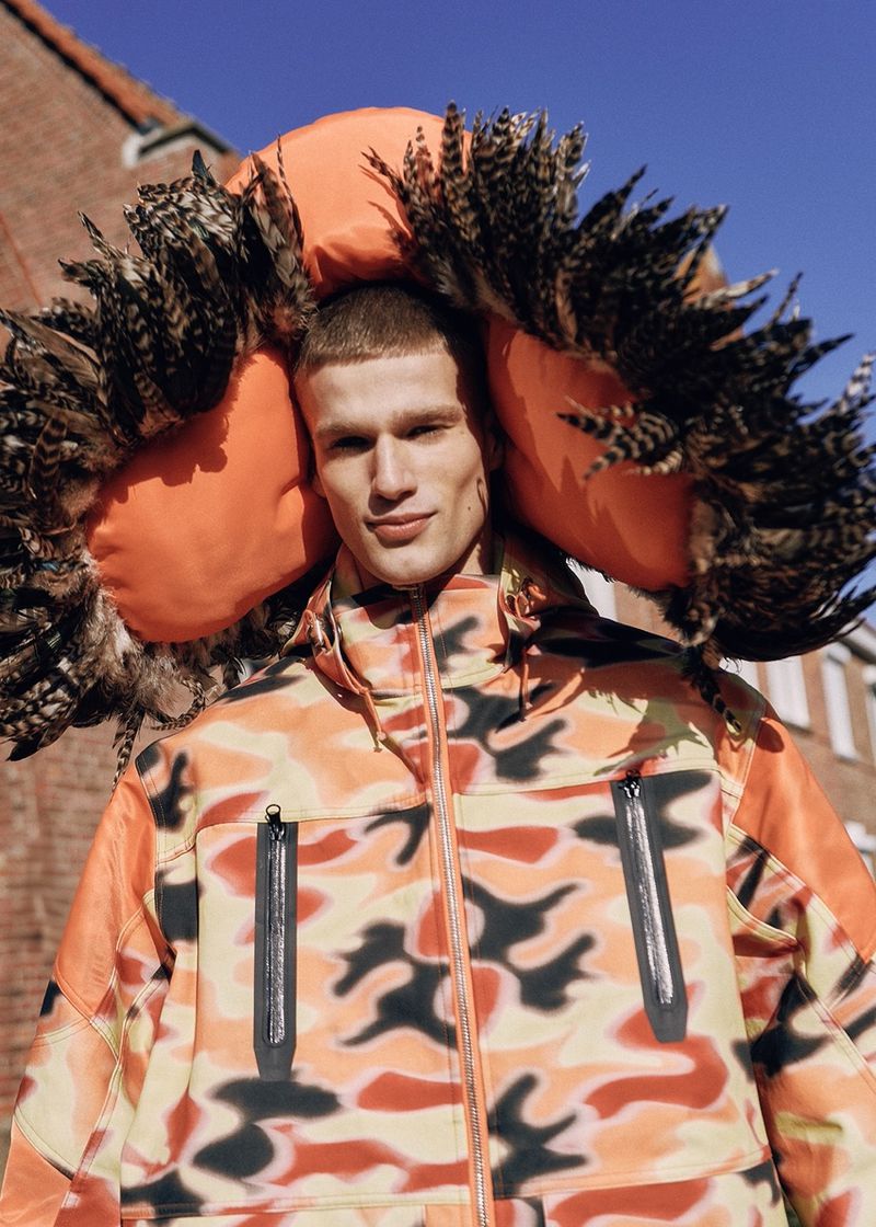 Portrait of a man in an orange camo jacket with a feather and puffy hood standing outside on a sunny day. Fashion editorial shot.