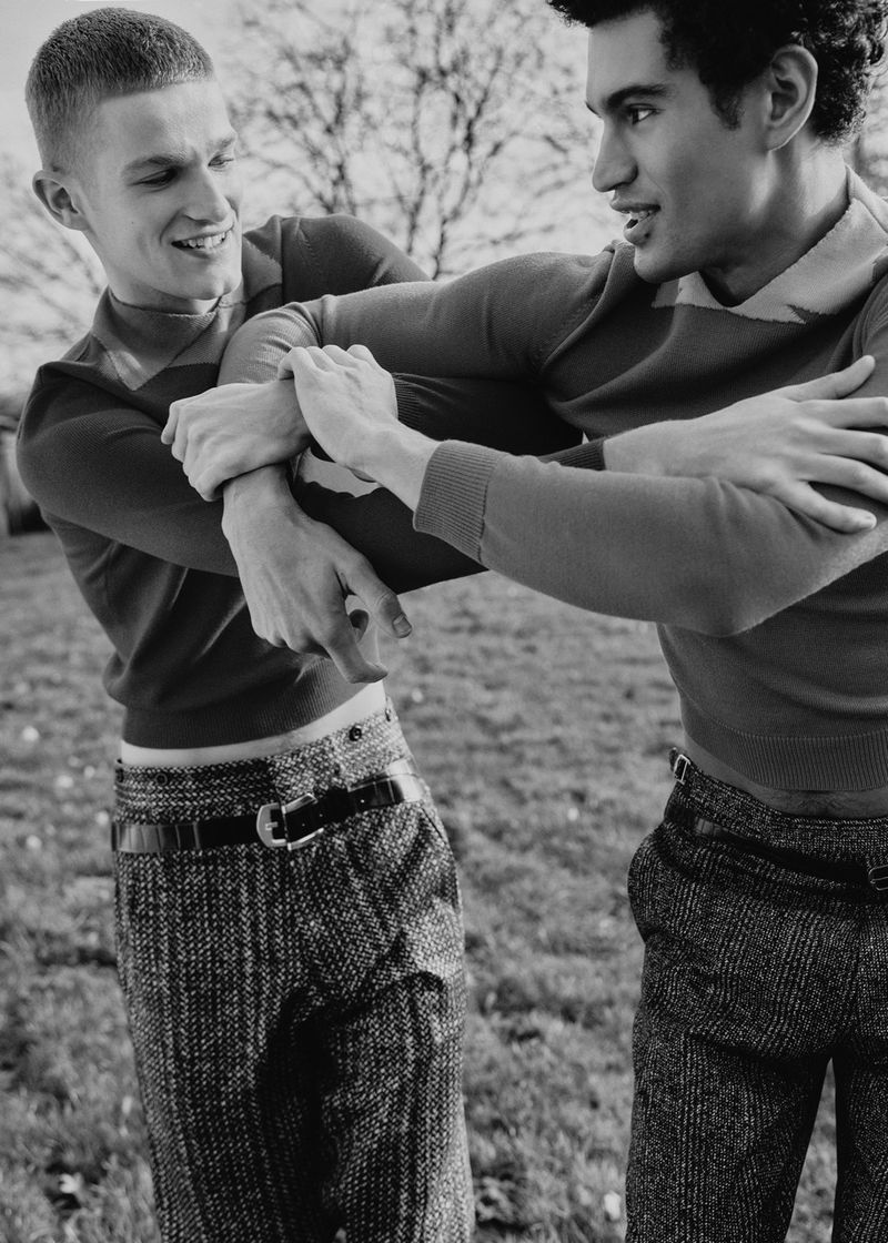 Two young adult men in fitted sweaters and patterned trousers playfully pushing each other in a grassy outdoor setting with bare trees.