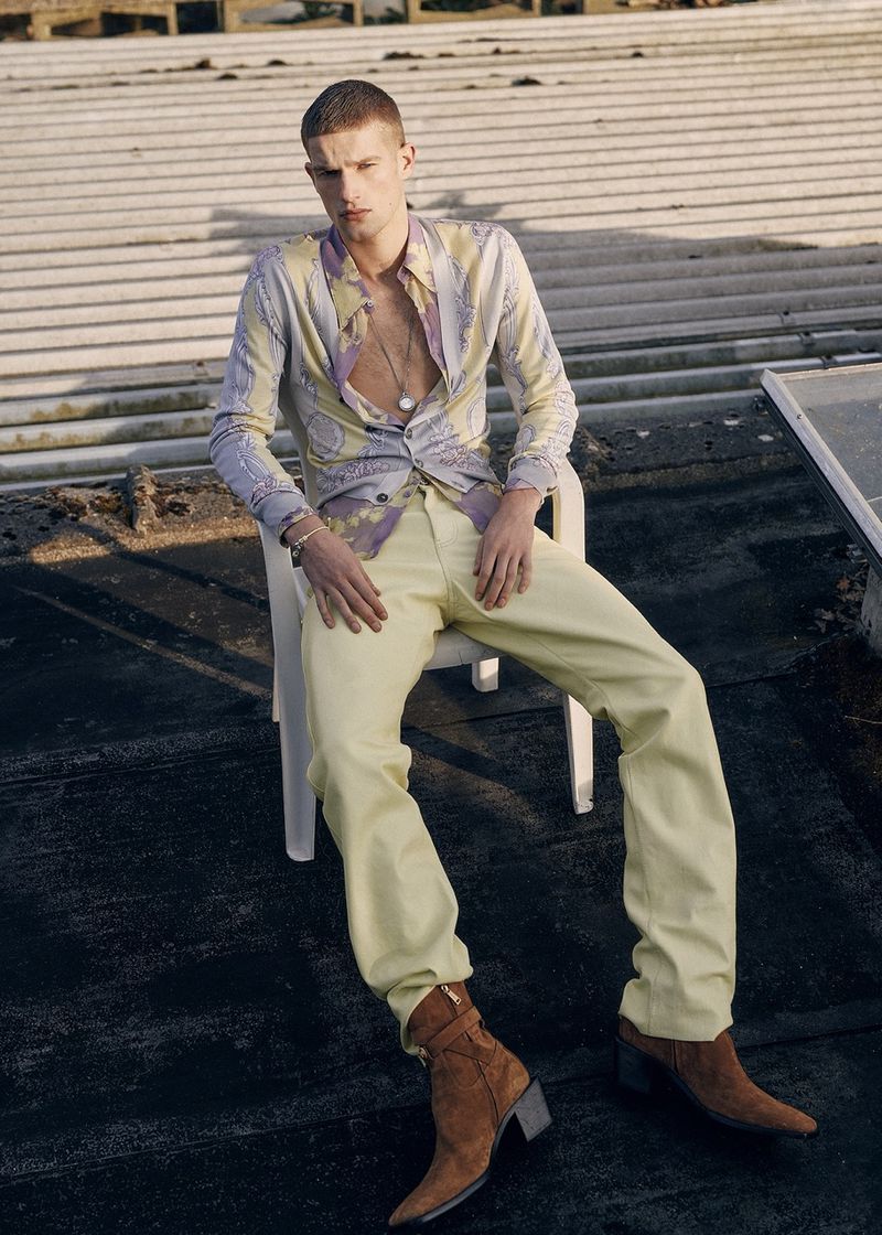 A slender, fair-skinned adult male model sits on a white chair outdoors, wearing an unbuttoned patterned shirt, yellow trousers, and brown cowboy boots.