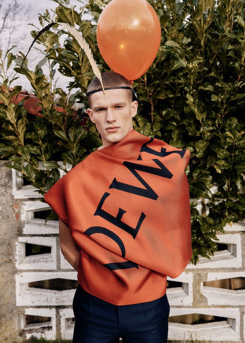 Man in navy pants and orange fabric with Loewe logo posing outdoors with an orange balloon in front of greenery and a white brick wall.